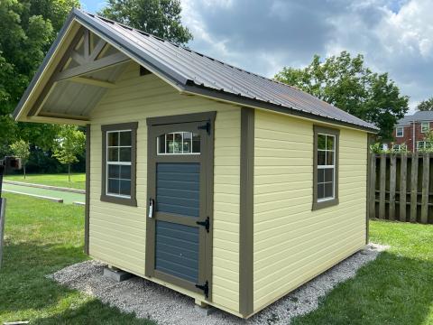 Yellow garden shed with green trim, a blue single door, multiple windows, door awning, and a metal roof that sits in a backyard and can be used as a kid's playhouse.
